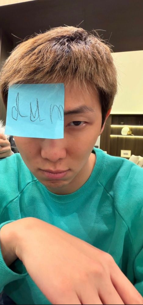 Namjoon wearing a turquoise crewneck sweater looking at the camera while a post it with the word “dum” on it is on his forehead