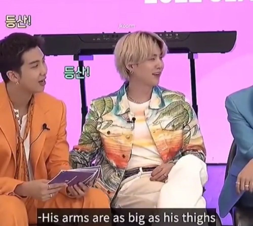 Blonde yoongi in a long sleeve kind of Hawaiian style shirt and white pants sitting next to Namjoon wearing an orange suit. Yoongi is having the time of his life talking about how big Namjoon is. The caption reads “his arms are as big as his thighs”