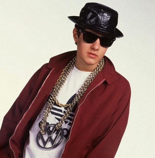 Photo of Mike D of the legendary Beastie Boys (circa 1986) wearing black leather bucket hat, black sunglasses, a white Adidas shirt, rust colored track suit top and long thick gold chains with giant VW logo pendant.