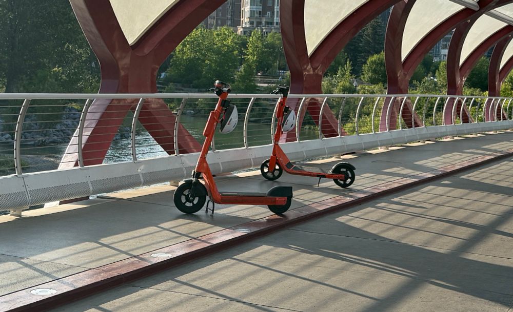 Two e-scooters, parked on a bridge so as to completely block the pedestrian path on one side.