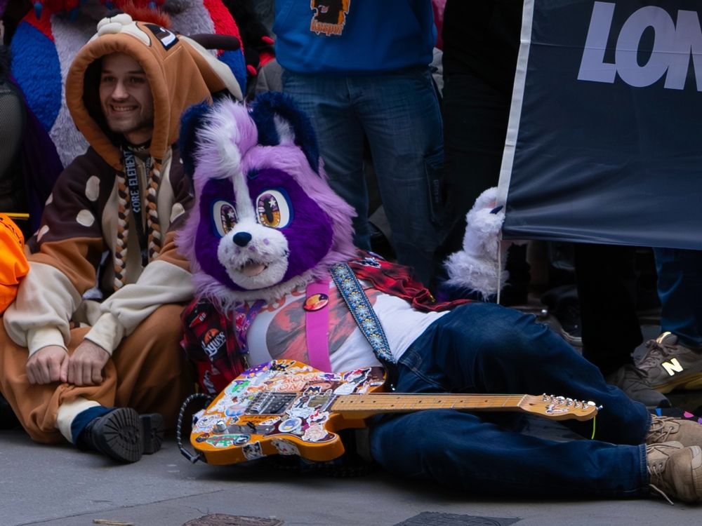 A photograph of Arcadia, a purple fox fursuiter, laying on the street with his guitar next to the LondonFurs banner.