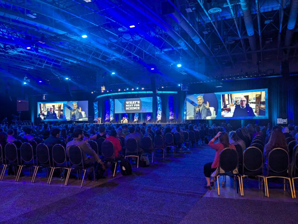 A colossal and cavernous room is bathed in blue light. On stage is a woman in a white chair with a big screen behind her that says WHATS NEXT FOR SCIENCE. Two absolutely ginormous screens, one to each side, project video of the woman in the chair and a Zoom feed with Gov Jay Inslee of Washington State. Hundreds of people are sitting in chair facing the stage.