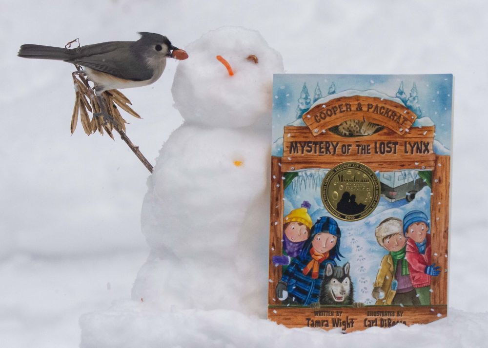 A tiny snowman beside the middle grade book, Lost Lynx. A tufted titmouse is on the snowman's arm.