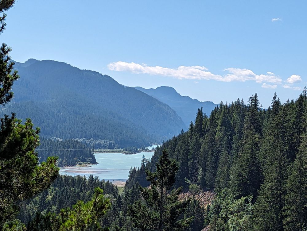 This image shows a body of light blue water framed by dark green fir trees. Large mountains are silhouetted in the background and the sky is blue with some thin white clouds.  