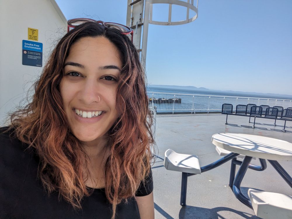 This image shows a selfie picture of Co-Investigator Sarah Huque on the sun deck of a ferry. Sarah is smiling at the camera and in the background, you can see plastic seats, a barrier and the ocean and mountains on the horizon.  