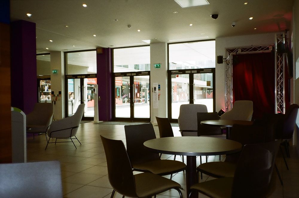 A film picture of an empty arts cafe. There are tables and chairs placed in front of a small stage to the right, and sun is streaming in through multiple glass doors to the back of the room.