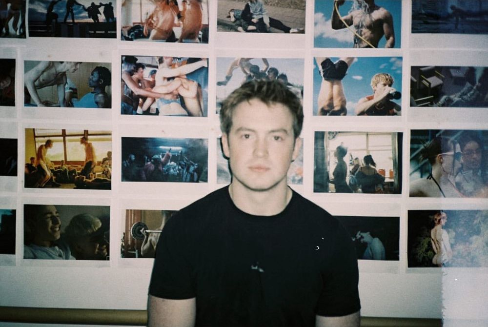 Kit Connor posing in a black shirt in front of pictures from Troye Sivan music videos. Looking fine as hell!