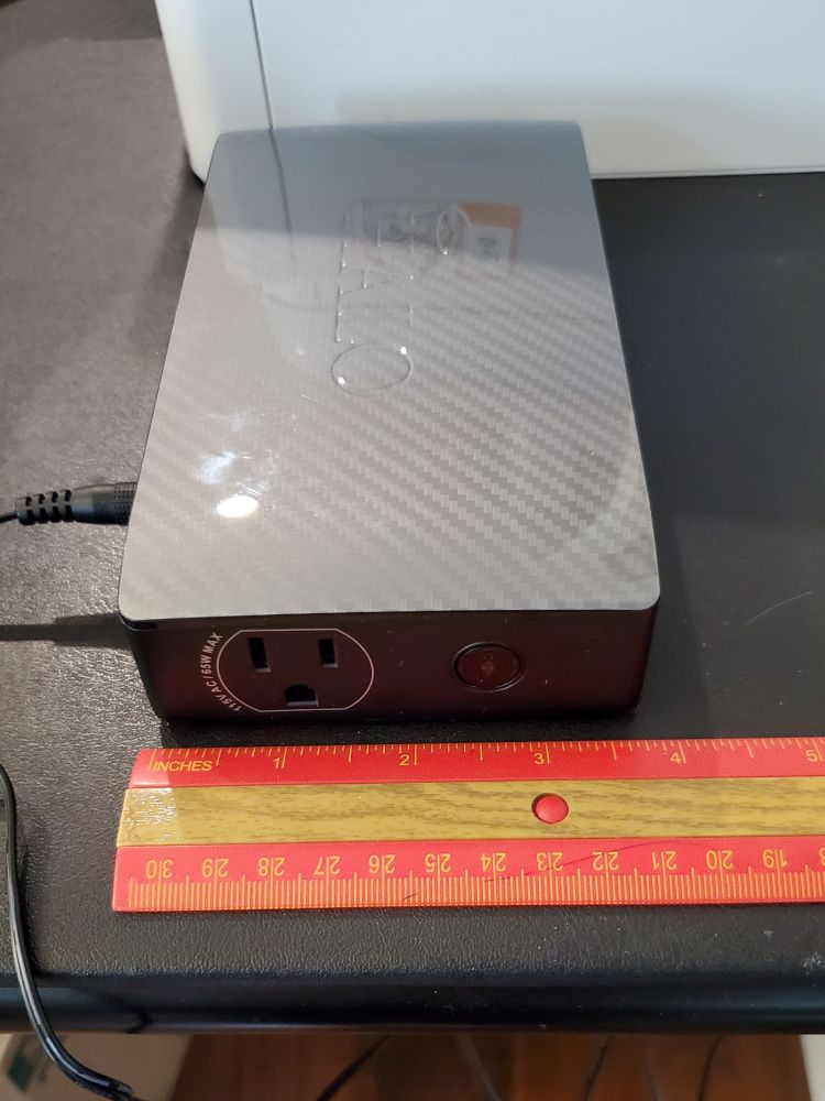 A HALO Bolt on a card table, rotated sideways so that the front faces to the viewer's left and the side with the 115V/65W outlet (similar to a standard AC wall outlet) is visible. An orange ruler lies on the table showing that the Bolt is about 3¾ inches wide on the short end.
The opposite end on the Bolt,  hidden from the viewer, has a floodlight that's 1¾ inches wide and ¾ inch tall.