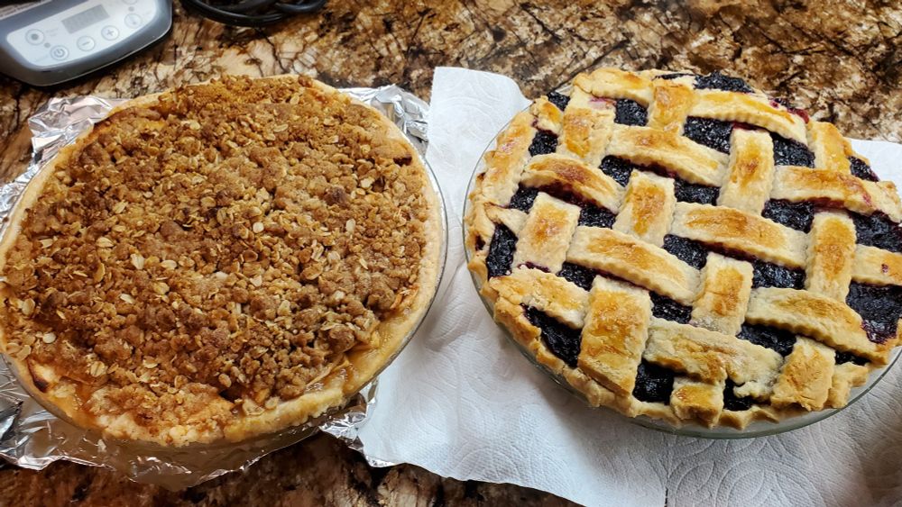 Overhead view of two homemade pies sitting on a kitchen counter. The pie on the left is a streusel-topped French apple pie, and the pie on the right is a triple berry pie with a lattice top crust.