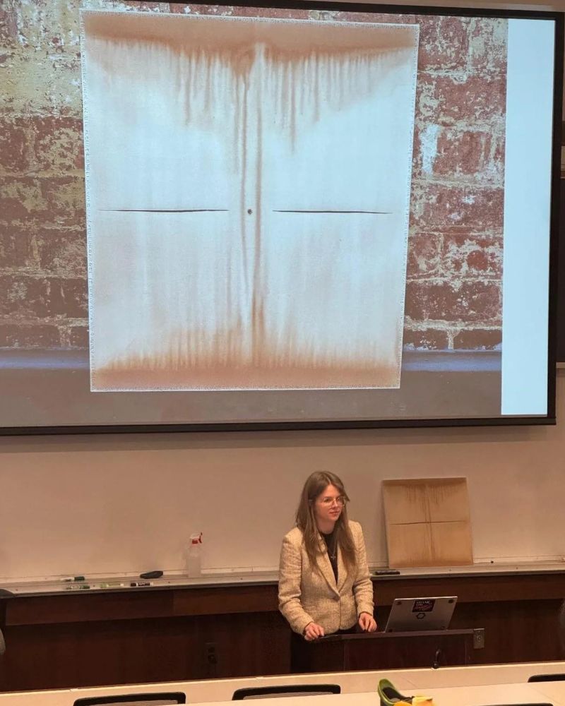 Me with a painting and a photo of the piece projected behind. It is an abstract piece with wine dripping below and above toward a slash and puncture in the center of the canvas 