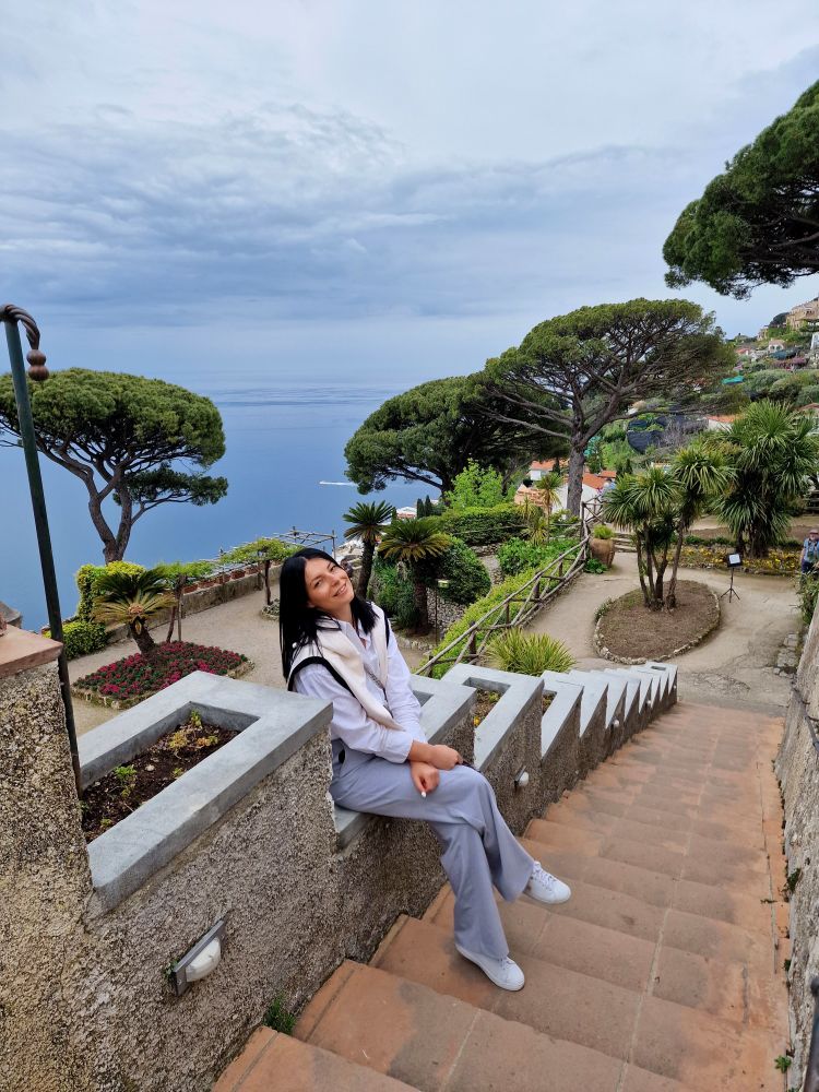 Sitting on a stone ledge at Villa Rufolo in Ravello, I smile at the camera, taking in the breathtaking view. 

The lush gardens, towering Mediterranean trees, and the endless blue sea below feel almost surreal.

The cloudy sky adds a dreamy touch to this magical place on the Amalfi Coast.