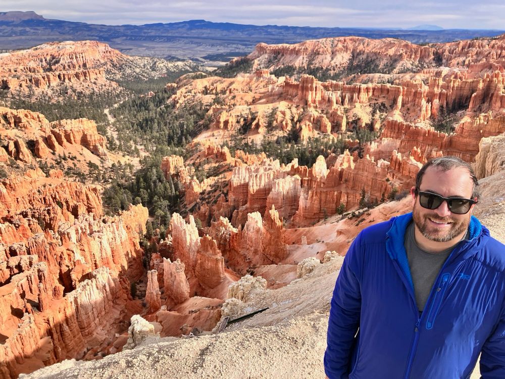 Eric standing with Bryce Canyon in the background. A warning sign has fallen over and down the ravine.