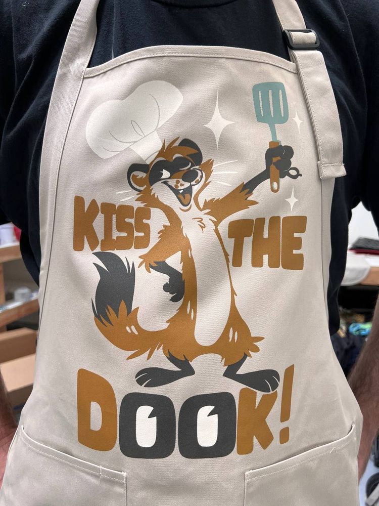 A cooking apron with a ferret on it and text that says "Kiss the Dook".