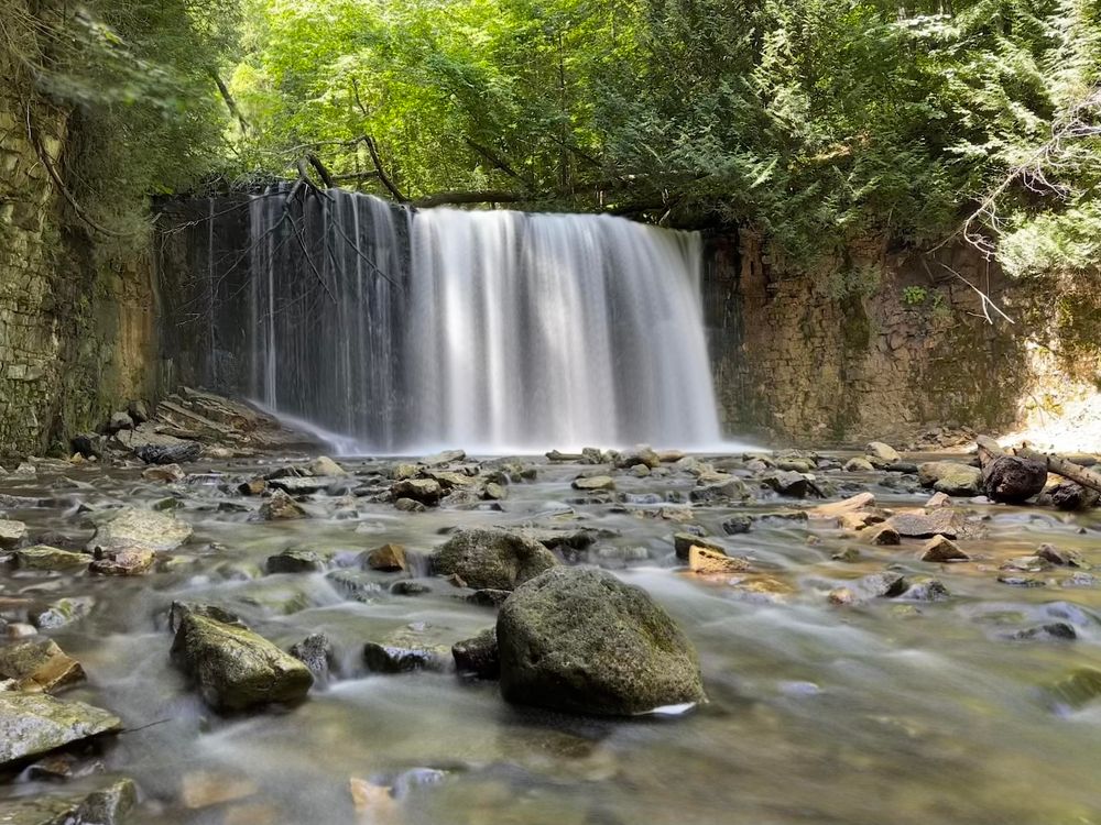 A block waterfall in a wooded area. Pic is taken from the lower level and shows that the waterfall plunges into a shallow but wide creek with lots of rocks.