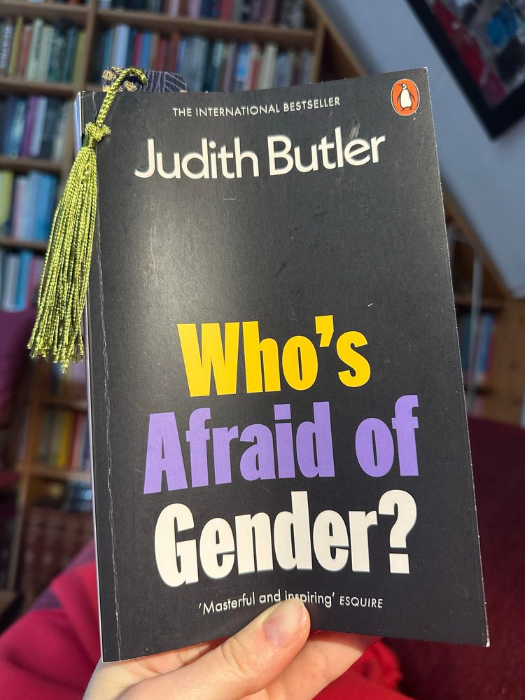 My hand holds the book. The cover features bold text in yellow, purple, and white against a black background. Cover describes it as an international bestseller, and as “Masterful and Inspiring”. A bookmark with a green tassel is stuck in the book. Background features a book case and part of a painting.