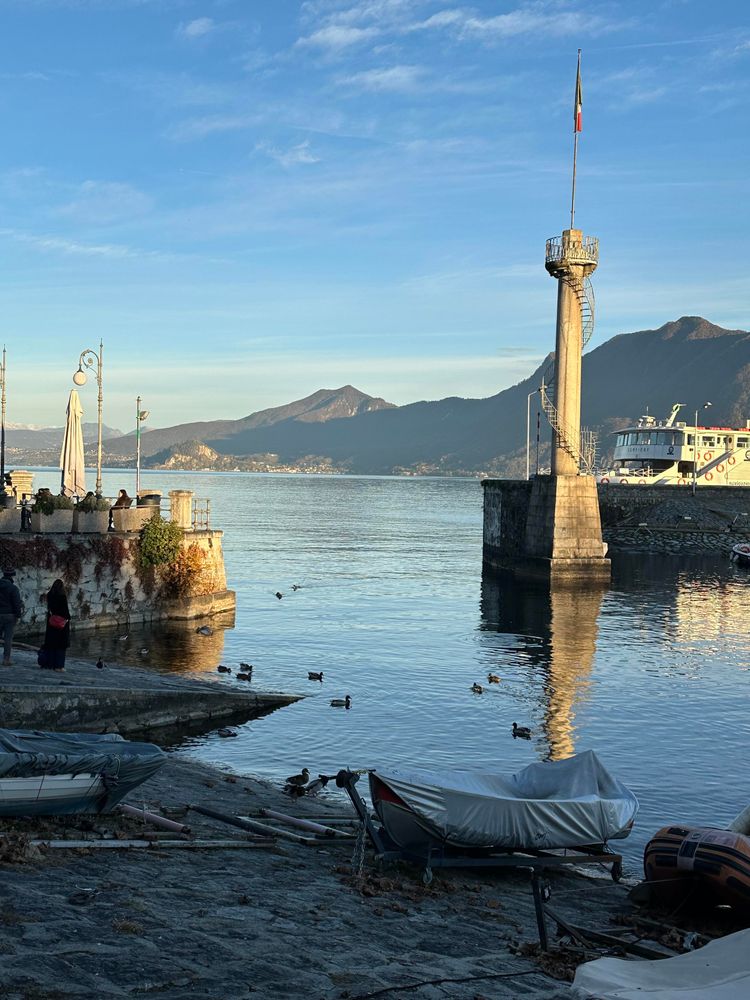 View of the exit of a harbour. A small concrete lighthouse-like thing with a spiral staircase around it and an Italian flag on top is located at the right side of the mouth of the harbour. In the foreground a few small boats. Behind the lighthouse a larger boat. Mountains in the background. Soft, golden evening light. A few ducks in the water. A patio with streetlights, tables and flowers on the left side of the harbour exit.