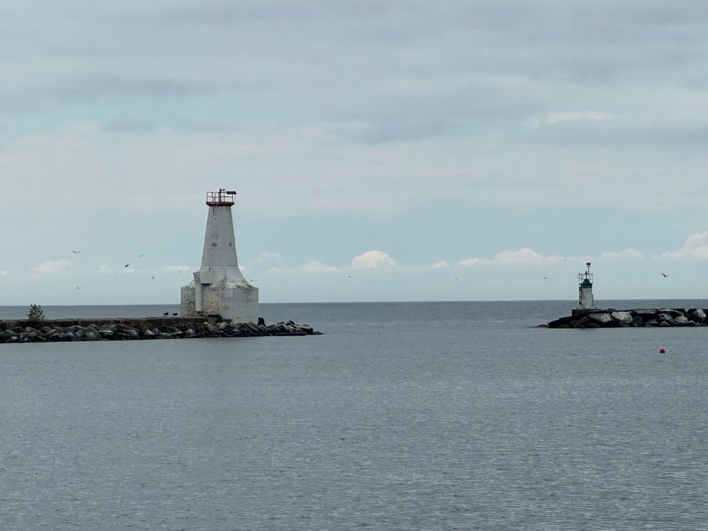 Exit from a harbour, seen from that harbour, looking out to the lake. Exit has a small lighthouse on the left and the end of a rocky pier and maybe a very small lighthouse on the right? Both are white. The sky is overcast with soft clouds, and there are birds flying.