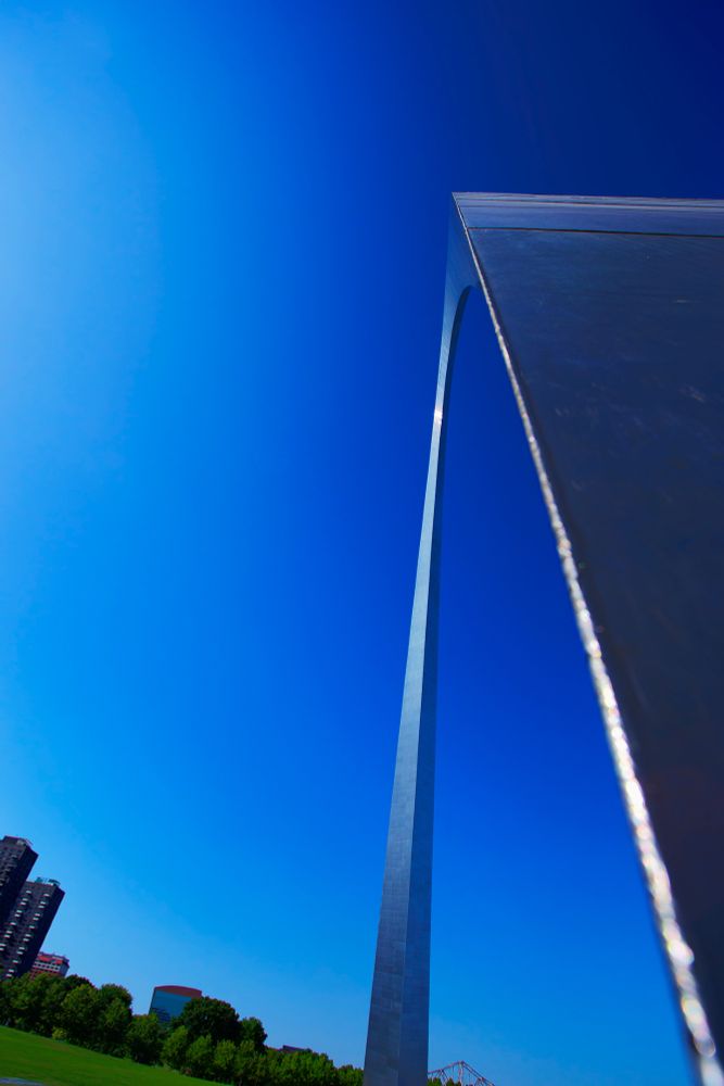 The bottom left of frame shows a corner of green grass and a row of trees fronting some buildings, the right side of frame shows the arc of a metal arch, but blue sky dominates most of the picture