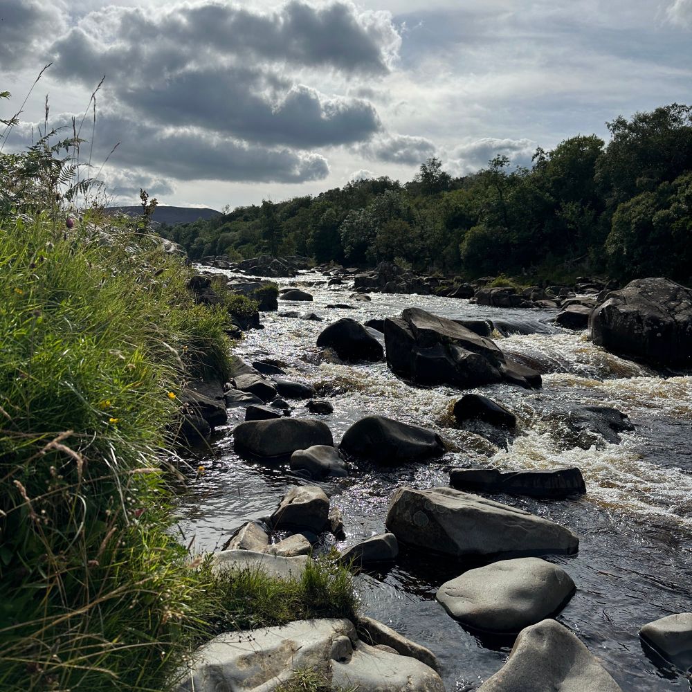 Looki by upstream along the Tees river, just above High Force. The water glimmers silver among black rocks and the banks are a lush green tumble. Moors and heavy cloud loom in the distance