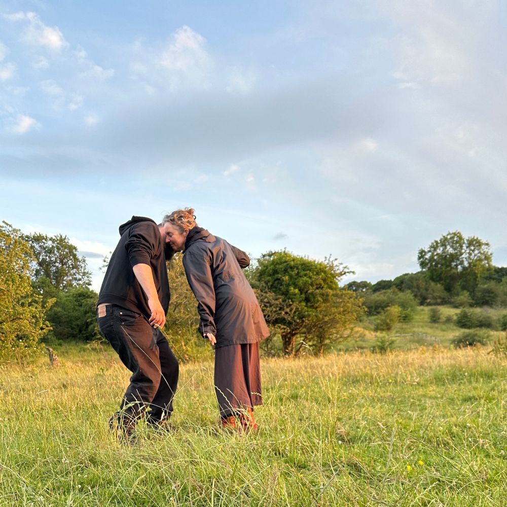 Two people practicing movement improvisation in a field. Their cheeks are pressing together and their movement revolves around the point of contact. There are hawthorn trees behind them and the grass glows in evening light 