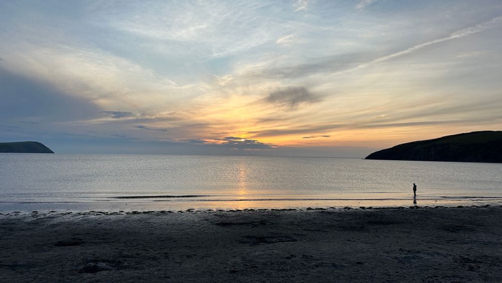Sunset over a silvery bay between steep headlands. A solitary figure stands silhouetted on the shoreline 