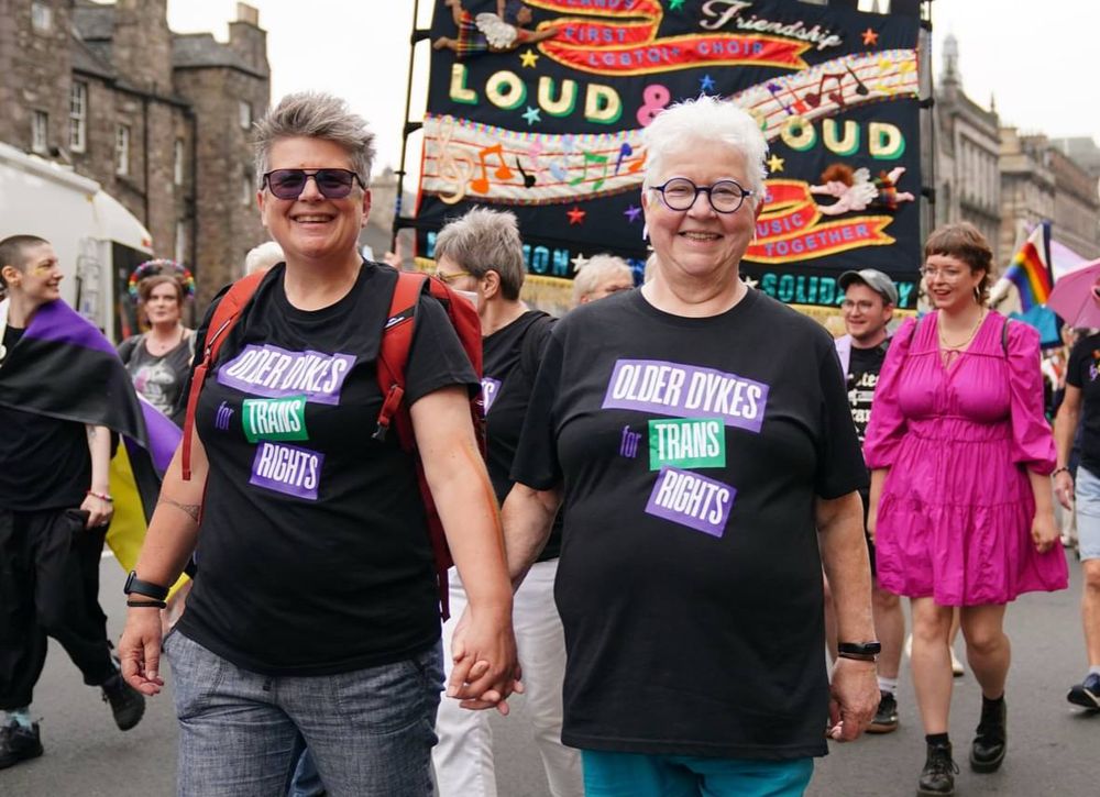 Photo of Val McDermid and Prof Jo Sharp at Edinburgh Pride march. They are wearing big grins and matching black T shirts that read ‘older dykes for trans rights’ in purple, green and white.