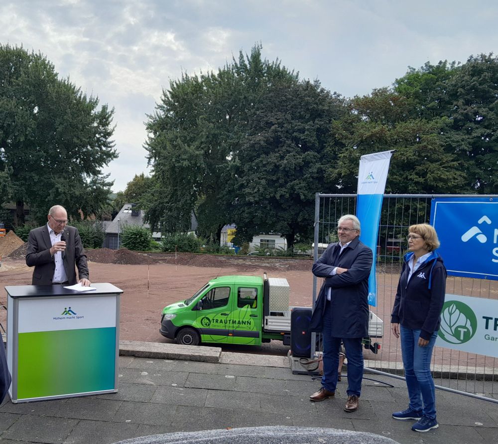 Der Bezirksbürgermeister hält am Pult eine Rede zum ersten Spatenstich, der Oberbürgermeister und die Leiterin des Mülheimer Sportservices hören zu.