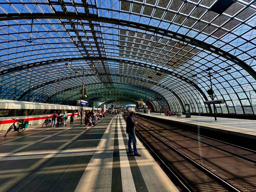 Arching roof over train tracks divided into a million squares as blue skies show through.  Random humans wait on the platform with sun streaking through.