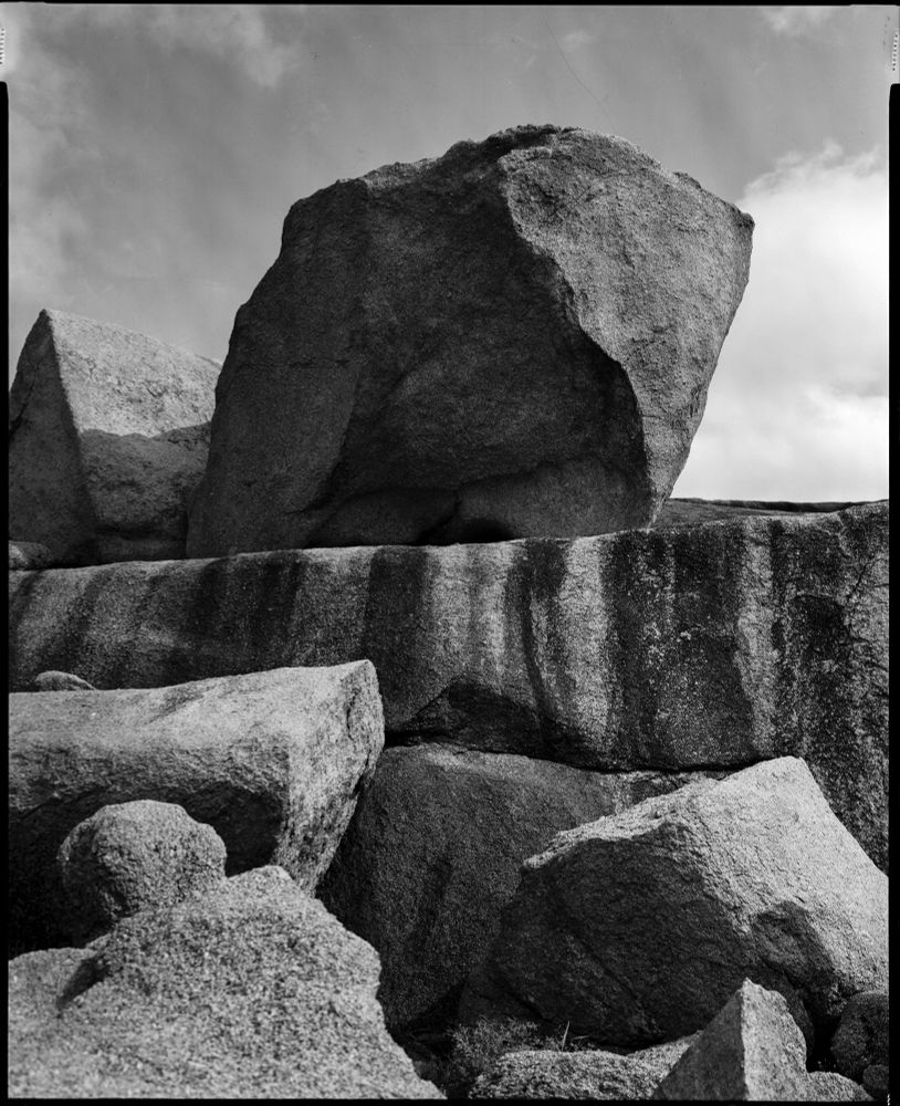 Black and white photograph of massive rock formations at Enchanted Rock, Texas, captured with an Intrepid 8x10 large format camera on Bergger Pancro film. The image emphasizes the rugged textures and imposing scale of the rocks under a partly cloudy sky, with strong contrasts highlighting the layered surfaces and natural erosion patterns.
By Thomas Hooper. 