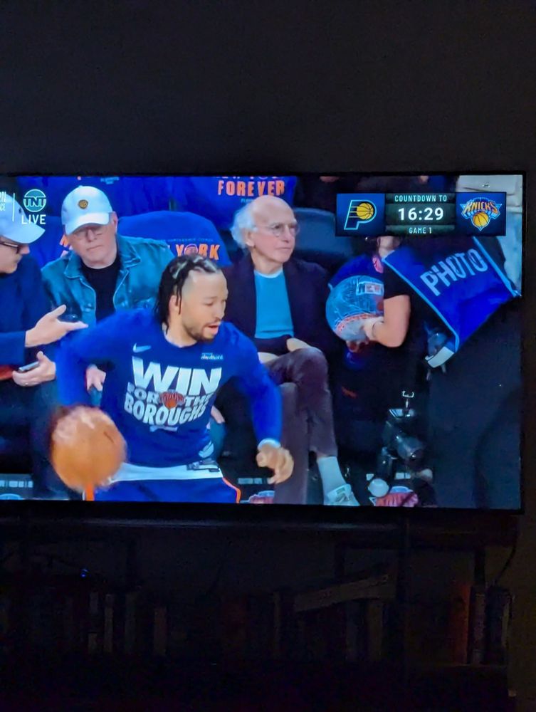 larry david sitting courtside behind new york knick jalen brunson at a basketball pregame warmup 