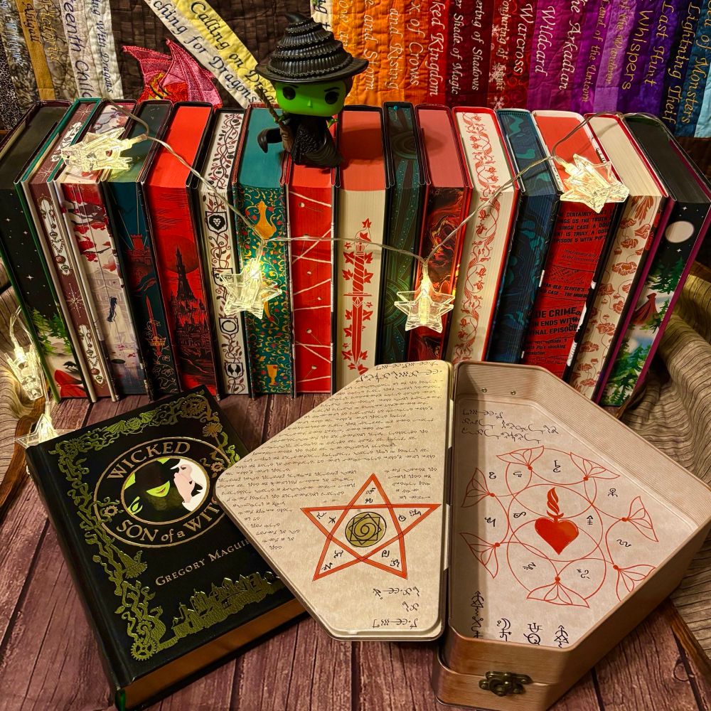 Open Grimmerie tin beside Barnes & Noble Leatherbound Edition of “Wicked” and “Son of a Witch” combined book, back by red and green stenciled edges and star lights with Elphaba Funko Pop on top