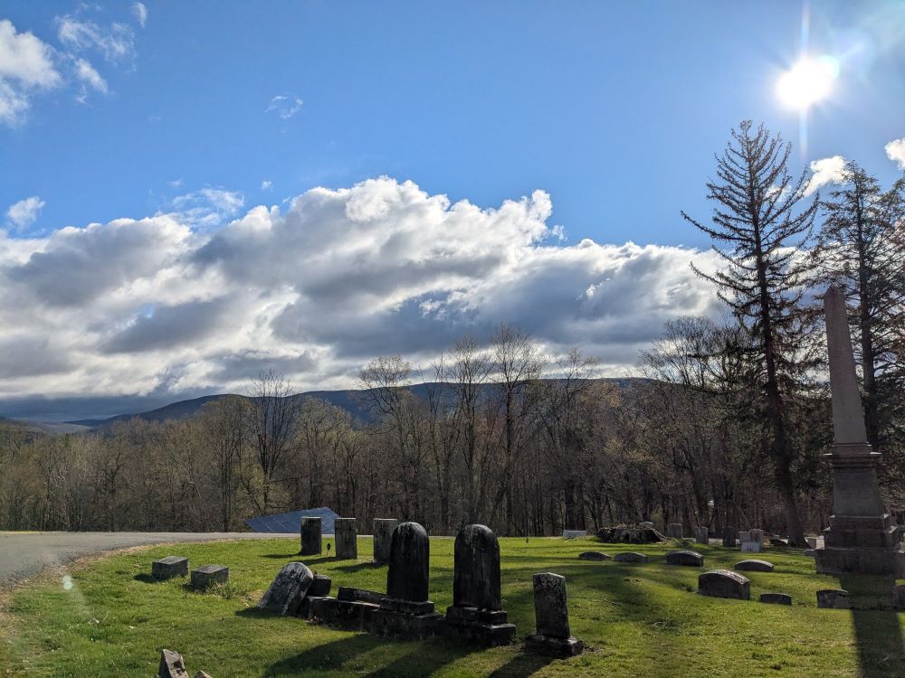Blue sky, a few clouds, and sun, over a green corner of a cemetery 
