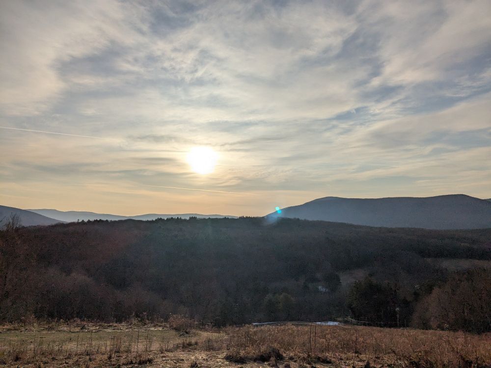 Bright yellow sun through filmy white clouds, glinting over mountains and a brown early spring field.