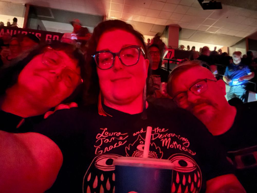 Three people cast in red, two on the sides have their heads on the middle one’s shoulders 