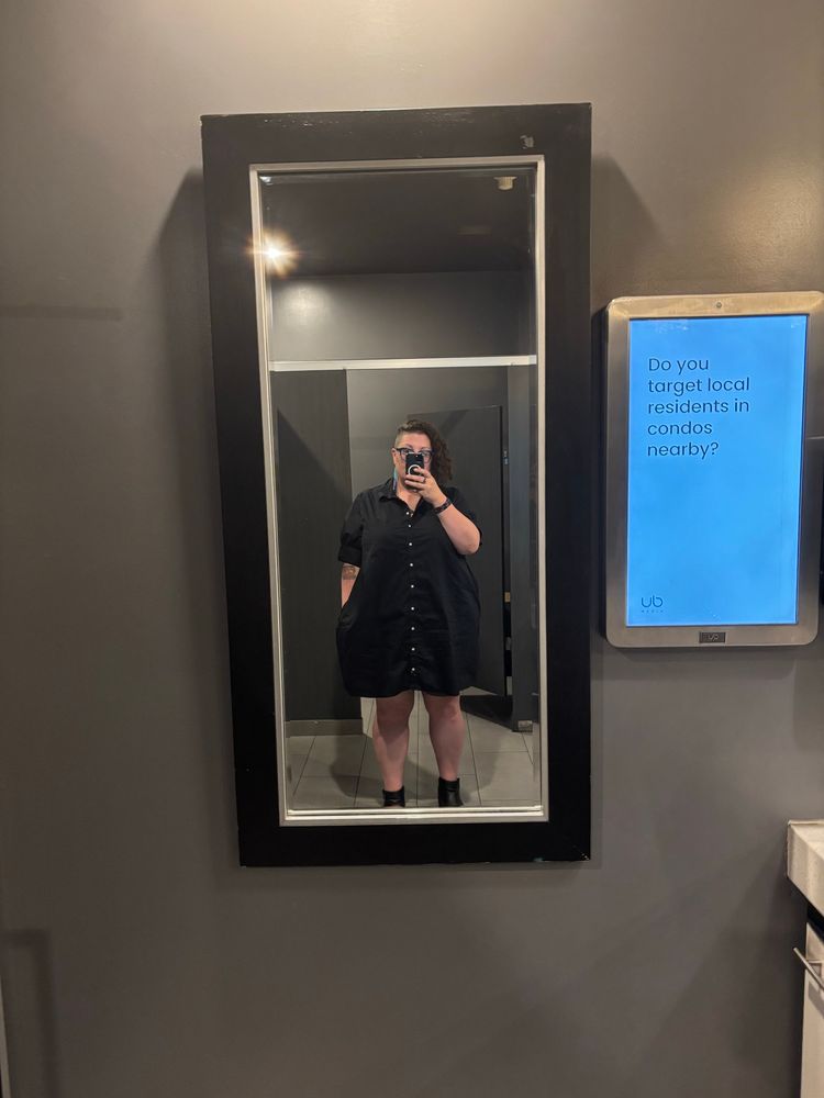 Mirror selfie of a woman wearing an oversized black “dress shirt” dress. Phone obscures her face. 