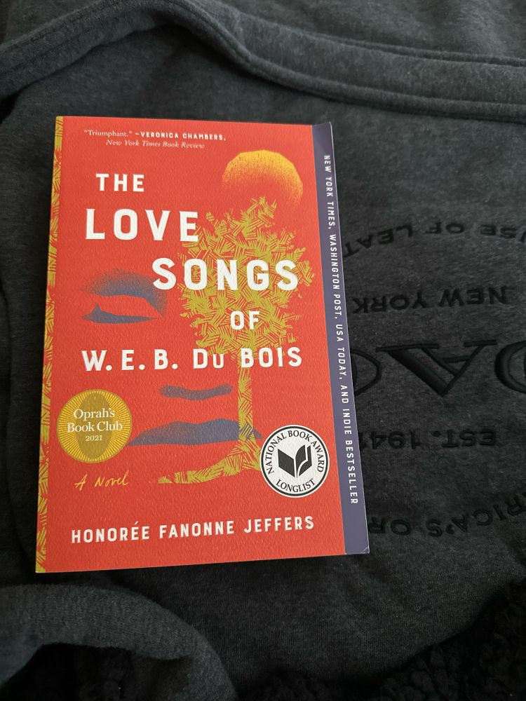 The image shows a copy of the book The Love Songs of W.E.B. Du Bois by Honorée Fanonne Jeffers. The book’s cover is red-orange with yellow and black accents, featuring abstract illustrations, and displays seals for Oprah’s Book Club 2021 and the National Book Award Longlist. It is resting on a dark gray Coach brand robe.