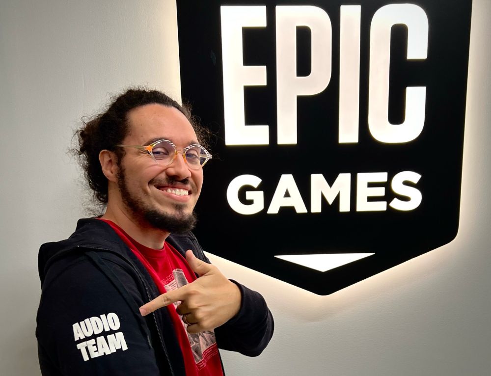 Me wearing a red shirt and a black hoodie with “AUDIO TEAM” in bold white capital letters. I’m at Epic Games HQ, standing next to a large black “EPIC GAMES” badge with white letters on a white wall.