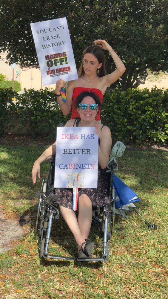 Woman in a wheelchair with a sign that says ‘IKEA HAS BETTER CABINETS’. A younger woman standing besides her with a sign that says ‘You can’t erase history. HANDS OFF’