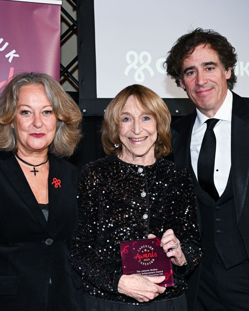 Three people smiling at an awards event. The person in the center is holding a plaque that reads "Carers Trust Awards 2023 The Johnnie Walker Lifetime Achievement Award." They are dressed formally, with dark attire and a backdrop featuring a logo.