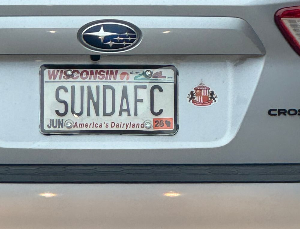 A closeup photo of a Wisconsin vanity plate on a Silver Subaru. It reads SUNDAFC and there is a Sunderland club crest sticker to its right.