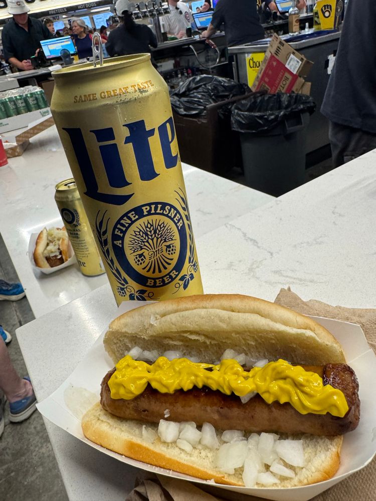 A brat on a bun with diced white onions beneath and a generous serving of yellow mustard across the top lengthwise. The mustard is oddly splotchy in its distribution because it was pumped onto the brat from a dispenser on the field level at American Family Field during a Brewers game. There is a 24 ounce can of Miller Lite immediately behind the brat.
