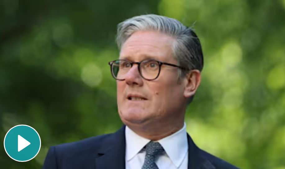 UK Prime Minister- Keir Starmer with grey hair wearing glasses, a dark suit, white shirt, and patterned tie stands outdoors in front of a background of green foliage.
