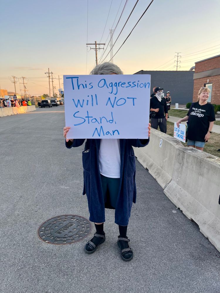 A guy dressed up as “The Dude” from The Big Lebowski holds a sign that reads “This aggression will not stand man.”