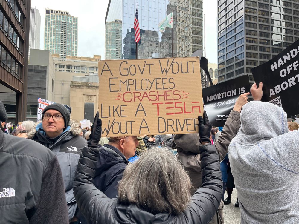 A person at a protest holds a sign that says “a government without employees crashes like a Tesla without a driver”