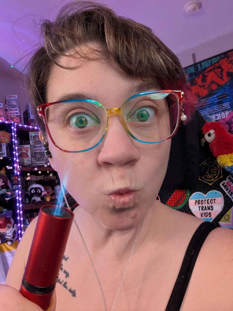 A person with short brown hair and rainbow glasses makes an "ooooo" face at the camera, while holding a lit, red butane lighter in front of their face
