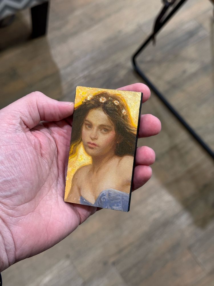 Small oil painting the size of a business card. Subject is a woman eating a lavender corset top. She has long brunette hair and is wearing a crown of small white flowers. The sky is a warm yellow. 