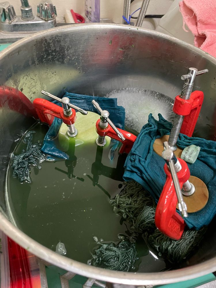 Silk scarves, folded multiple times and clamped to wooden disks that are acting as resists for the dye. The clamped scarves are in a pot of murky green water with yarn and a block of ice. 