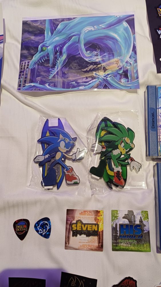 A small print of Super Sonic vs Perfect Chaos, 2 plastic standees (Sonic, Jet), 2 guitar pics with sonic logo on it and live and learn in the style of sonic Adventure 2 logo. Seven Rings in Hand pin badge in the style of Sonic and the Secret Rings and His World pin badge in the style of Sonic 06 logo. 