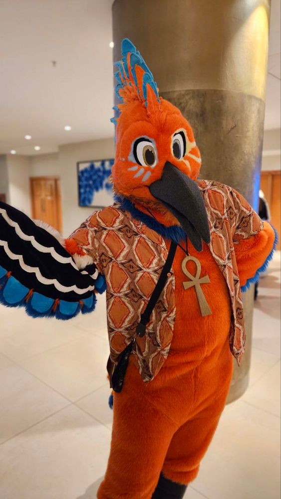 Photo of a orange and blue highlighted bird wearing a orange patterned shirt wearing an ankh necklace posing with one arm out and the other on his waist. 