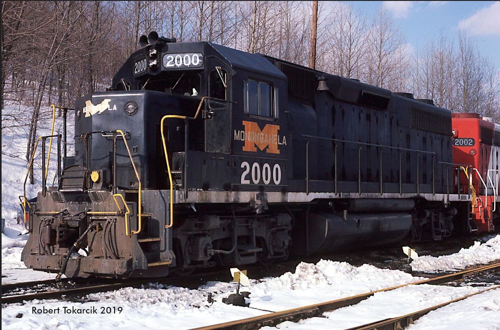 A black diesel locomotive with an orange M on the side  and the number 200, with red white and (out of frame) blue #2002 behind it.